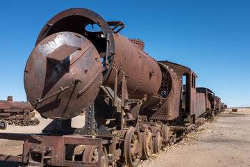 Fototapeta na wymiar A view along the side of a rusting steam train and carriages slowly rot away at the train graveyard just outside of Uyuni, Bolivia, against a bright blue sky.