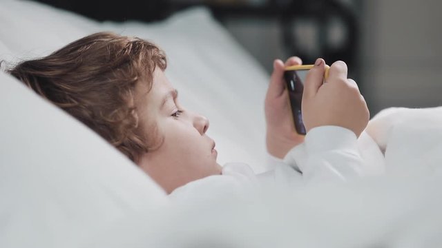 Little boy lying in the bed watching cartoon at modern yellow smartphone with interest.