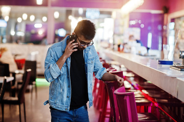 Stylish asian man wear on jeans jacket and glasses posed against bar in club and speaking on mobile phone.