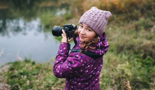 A young girl in a purple jacket and hat walks with a camera on the nature in the fall. Baby photographer. Happy childhood. Portrait and photography.