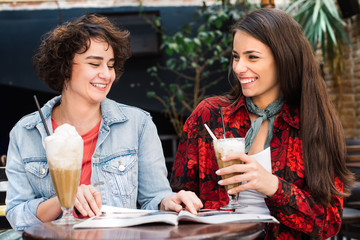 Hipster girls reading magazine at the bar. Girlfriends having a coffee and reading a newspapers at coffee shop. Beautiful girls drinking coffee having a conversation