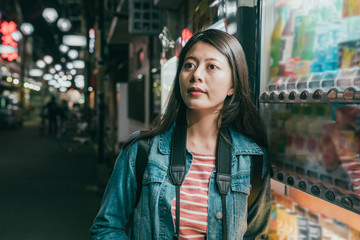 elegant curious young asian woman stand next to vendor machine in Japan late night city street. girl traveler standing in dark urban osaka japan looking aside not interested in vending drink auto.
