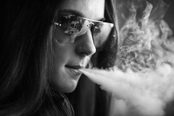Fototapeta premium Vape teenager with problem skin. Portrait of young cute girl in sunglasses smoking an electronic cigarette in the bar. Bad habit that is harmful to health. Vaping activity. Black nad white.