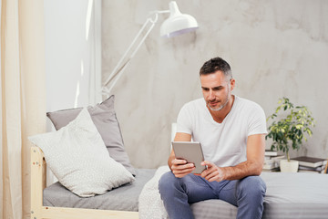 Caucasian man in forties sitting on bed in bedroom and using tablet.