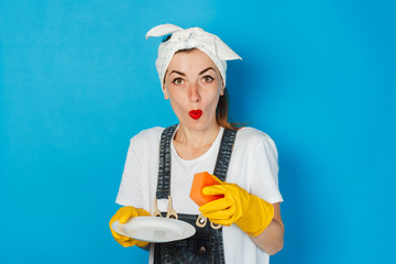 Young girl with a scarf on her head and yellow rubber gloves holds a white plate and a sponge on a blue background. Concept of cleaning and cleaning service, washing dishes.
