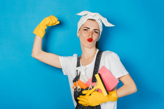 A young girl with a scarf on her head, yellow rubber gloves shows muscle biceps on her arm against a blue background. The concept of cleaning and cleaning service, high quality.