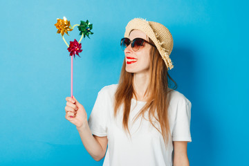 A young girl in glasses and a straw hat is holding a pinwheel toy, a blue background. Concept vacation, tourist trip, summer and tropics, summertime.