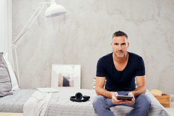 Caucasian man in forties sitting on bed in bedroom and using tablet.