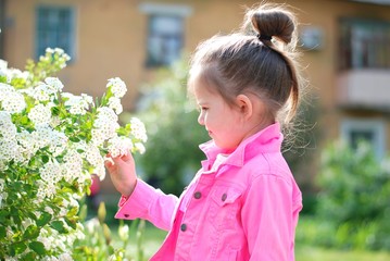 Little caucasian girl with bundled hair in a neon pink jacket holding blooming branch with white beautiful flower at the sunny summer day. Cute toddler girl smelling a white flowers on a blossom bush