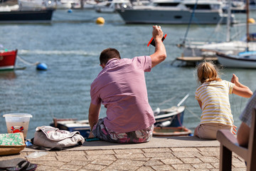 A father and daughter crabbing over the side of a harbour in the summer with sailboats in the background