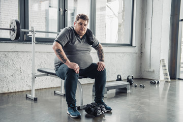 tired Obese man sitting on bench and Looking At Camera after exercising with dumbbells at gym - Powered by Adobe