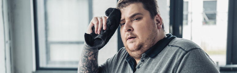panoramic shot of tired overweight man looking at camera and wiping face with towel at sports center