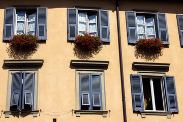 Beautiful charming windows with shutters on a light brown facade of residential building with green balcony and flowers on it in Bergamo Italy.