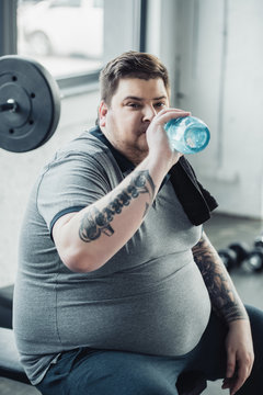 Tired Overweight Tattooed Man Drinking Water From Sport Bottle At Gym