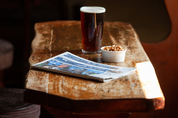 A pint of beer/ale in a pub on a wooden table with a newspaper and a ramekin of nuts
