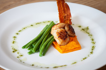 Freshly cooked scallops on a bed of sweet potatoes served with green beans, crispy bacon and salsa verde on a white plate in a restaurant