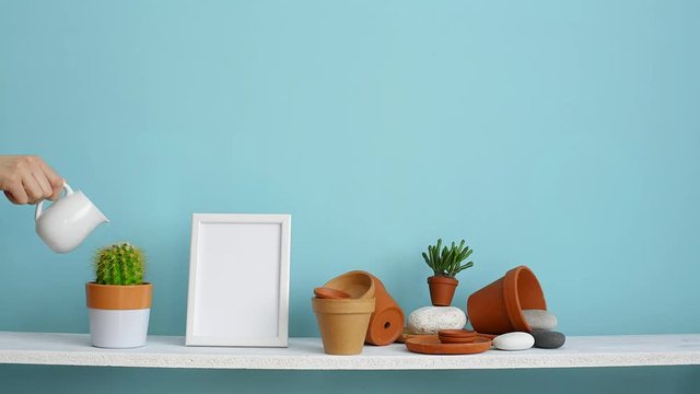 Modern room decoration with picture frame mockup. White shelf against pastel turquoise wall with pottery and succulent plant. Hand watering potted cactus plant.