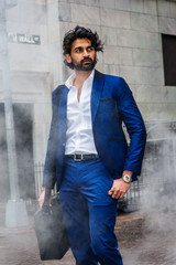 Obraz premium Raining day - grainy, foggy, wet feel. Young East Indian American Man with beard traveling in N, wearing blue suit, white shirt, carrying shoulder bag, looking away, walking on Wall Street in New York