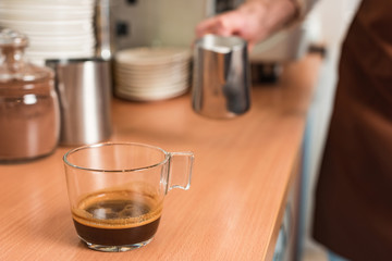 Cropped view of barista with milk jug and cup of coffee on wooden table