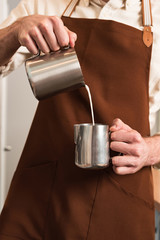 Cropped view of barista in brown apron pouring milk in steel jug