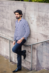 Raining day - grainy, drizzling, wet feel. Young East Indian American Man with beard, wearing black, white striped shirt, black pants, leather shoes, standing against wall outside in New York, thinks