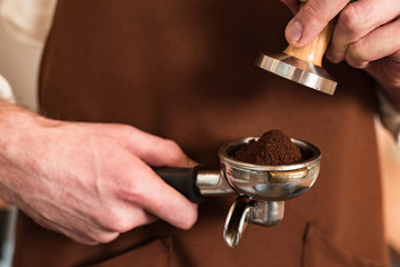 Cropped view of barista in brown apron holding portafilter with ground coffee and tamper