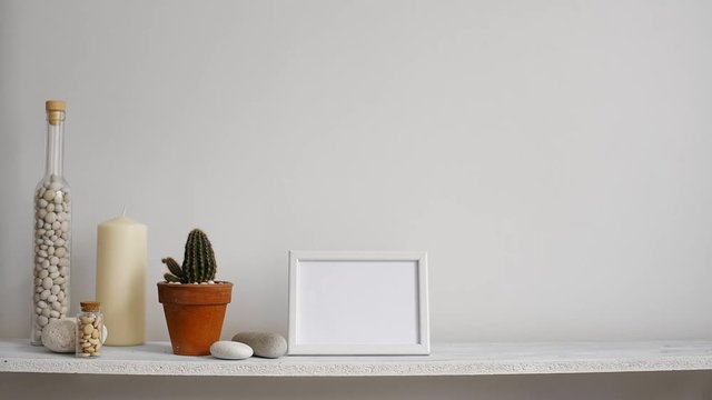 Modern room decoration with picture frame mockup. Shelf against white wall with decorative candle, glass and rocks. Hand putting down potted cactus plant.