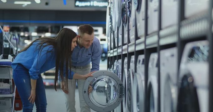 Young married couple man and woman in casual clothes appliances store choose to buy washing machine for home. Open the door looking into the drum, compare the design and characteristics of the devices