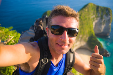 young happy and attractive backpacker man taking selfie portrait photo with mobile phone in front of amazing sea landscape smiling cheerful trekking on beautiful beach cliff