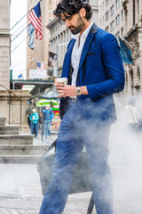 Raining day - grainy, foggy, wet feel. Young East Indian American Business Man with beard, wearing blue suit, white shirt, carrying leather hand bag, holding coffee cup, walking on street in New York