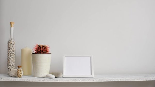Modern room decoration with picture frame mockup. Shelf against white wall with decorative candle, glass and rocks. Hand putting down potted cactus plant.