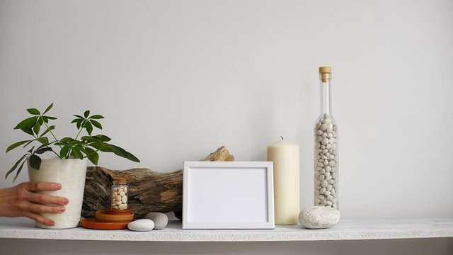 Modern room decoration with picture frame mockup. Shelf against white wall with decorative candle, glass and rocks. Hand putting down potted schefflera plant.