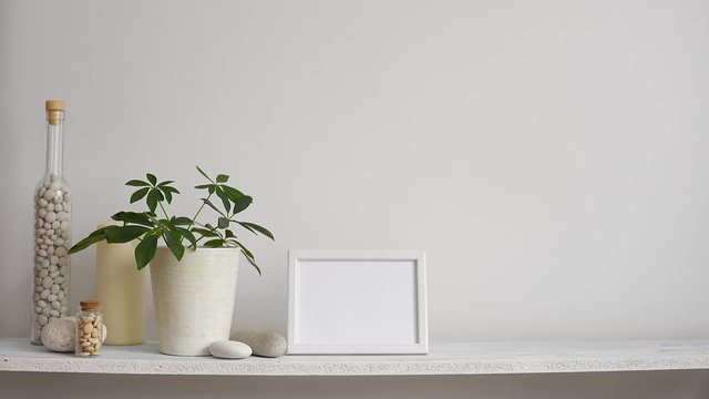 Modern room decoration with picture frame mockup. Shelf against white wall with decorative candle, glass and rocks. Hand putting down potted schefflera plant.