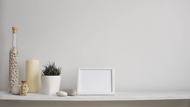 Modern room decoration with picture frame mockup. Shelf against white wall with decorative candle, glass and rocks. Hand putting down potted succulent plant.