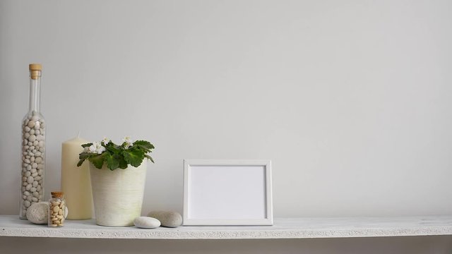 Modern room decoration with picture frame mockup. Shelf against white wall with decorative candle, glass and rocks. Hand putting down potted violet plant.