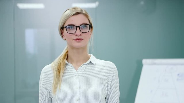 Young blonde businesswoman in office clothes and glasses is looking at camera and smiling.