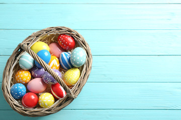Wicker basket with painted Easter eggs on wooden background, top view. Space for text