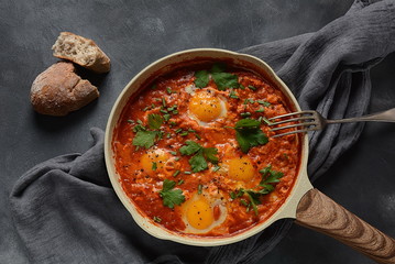 Shakshuka , middle eastern traditional homemade breakfast- fried eggs, onion, bell pepper, tomatoes and parsley in a pan on a dark background