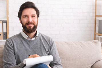 Bearded male psychotherapist writing on clipboard and smiling