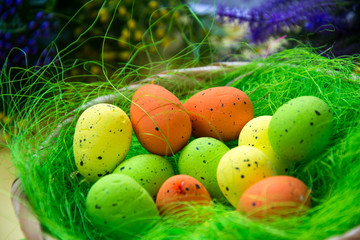 Fototapeta na wymiar Green nest in a basket with small colorful Easter eggs, decoration, close-up easter concept, holiday tradition, blurred paints and plastic flowers on yellow background, selective focus