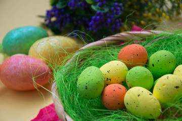 Green nest in a basket with small colorful Easter eggs, decoration, close-up easter concept, holiday tradition, blurred paints and plastic flowers on yellow background, selective focus