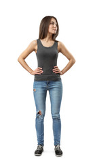 Front view of young attractive woman in gray sleeveless top and blue jeans standing and looking away with hands on hips isolated on white background.