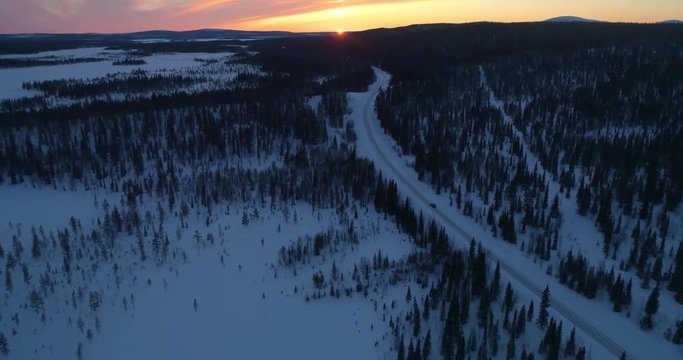 drone shot, following a vehicle, on a frozen route, between finnish woods and polar wilderness, in Pallas-yllastunturi national park, at the arctic circle, at sunset, on a sunny, winter evening dus