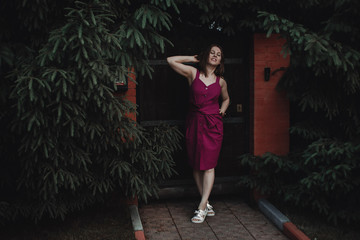 Stylish interesting girl in a Burgundy dress against a wooden fence and a thick hedge of fir trees....