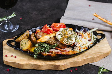 Hot assorted meats, bacon, meat sausages with roasted tomatoes, potatoes, poached egg and fresh cabbage on a platter, with a glass of red wine on a dark background