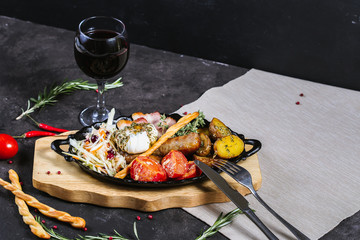 Hot assorted meats, bacon, meat sausages with roasted tomatoes, potatoes, poached egg and fresh cabbage on a platter, with a glass of red wine on a dark background