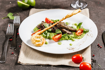Traditional German fried pork cheeks with greens and tomato closeup on a white plate with bread cakes on a served table with a glass of red wine
