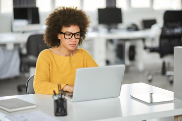Serious mixed race businesswoman dressed casual sitting in modern office and using laptop.