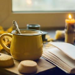 Fototapeta na wymiar Yellow mug with autumn leaves, a book and candles. The concept of comfort and warmth. Square framing.