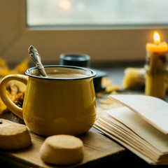 Yellow mug with autumn leaves, a book and candles. The concept of comfort and warmth. Square framing.